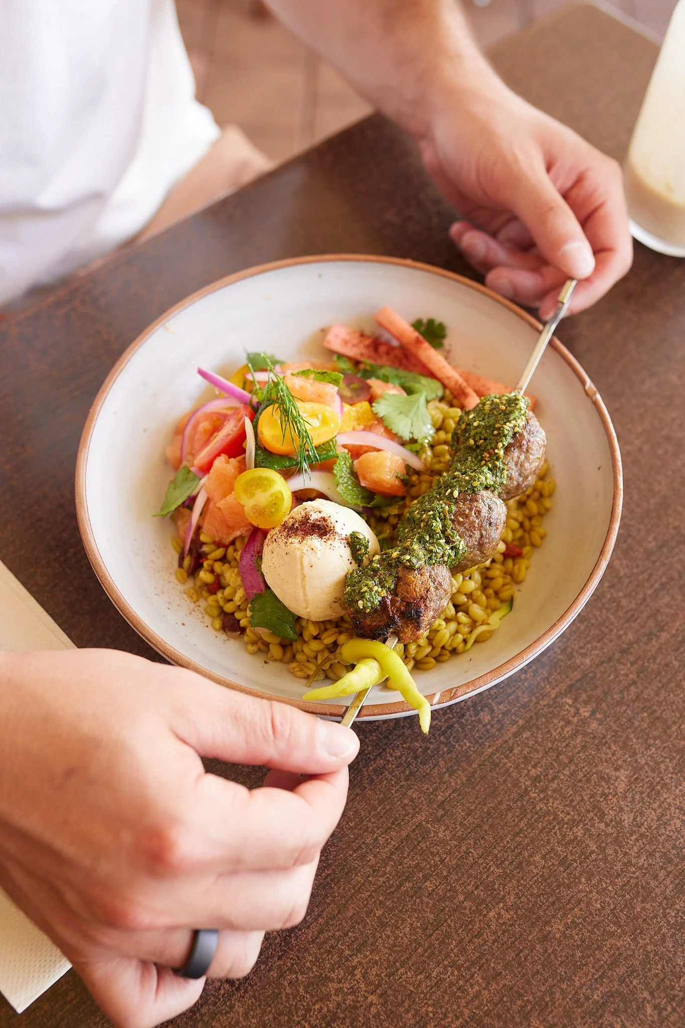 A person holding a bowl of colorful food, including vegetables, grains, and meat balls, with a person stirring the food with a fork.