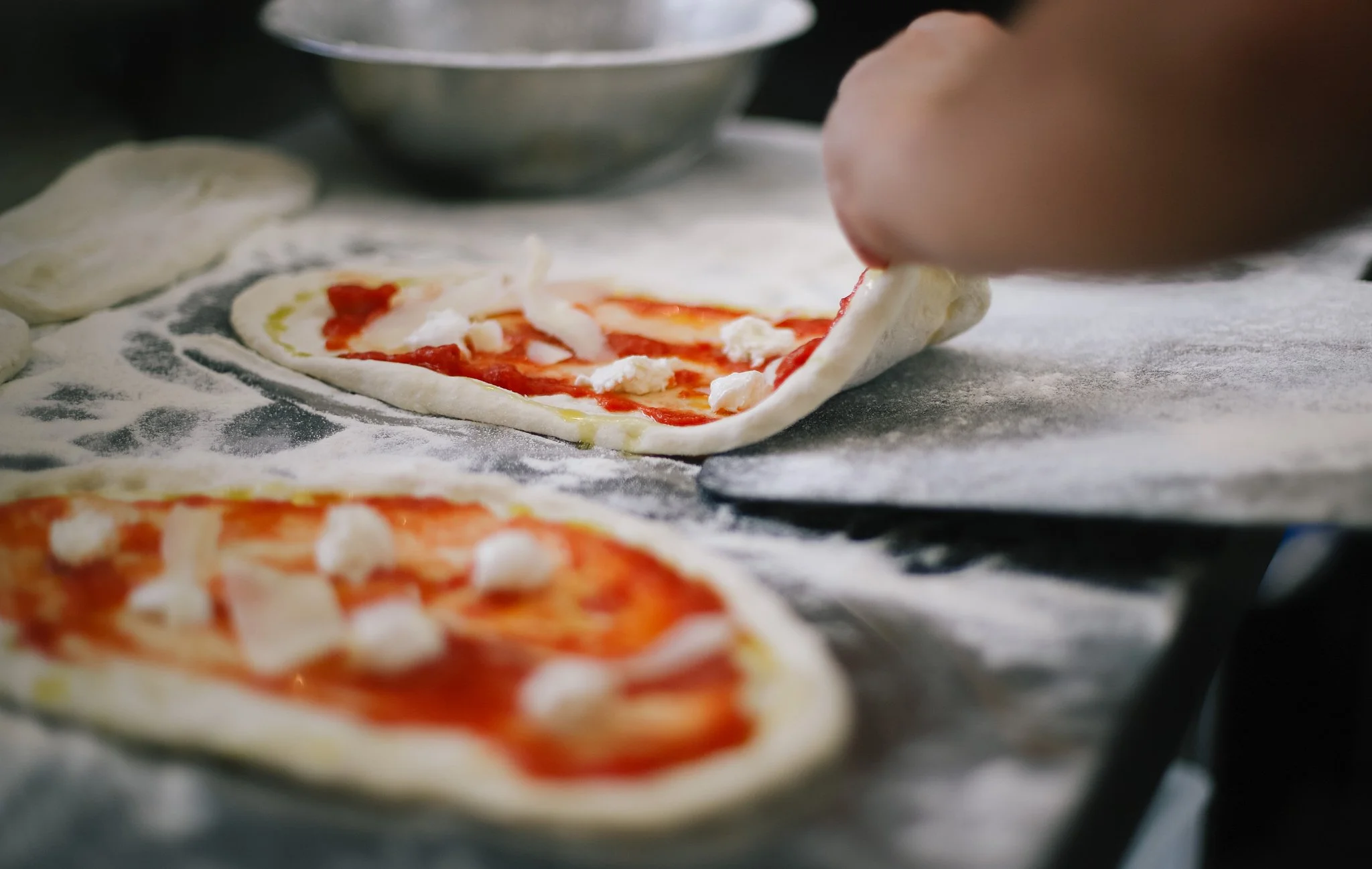 A person placing toppings on a small pizza with cheese and tomato sauce on a baking tray, with others in the background.