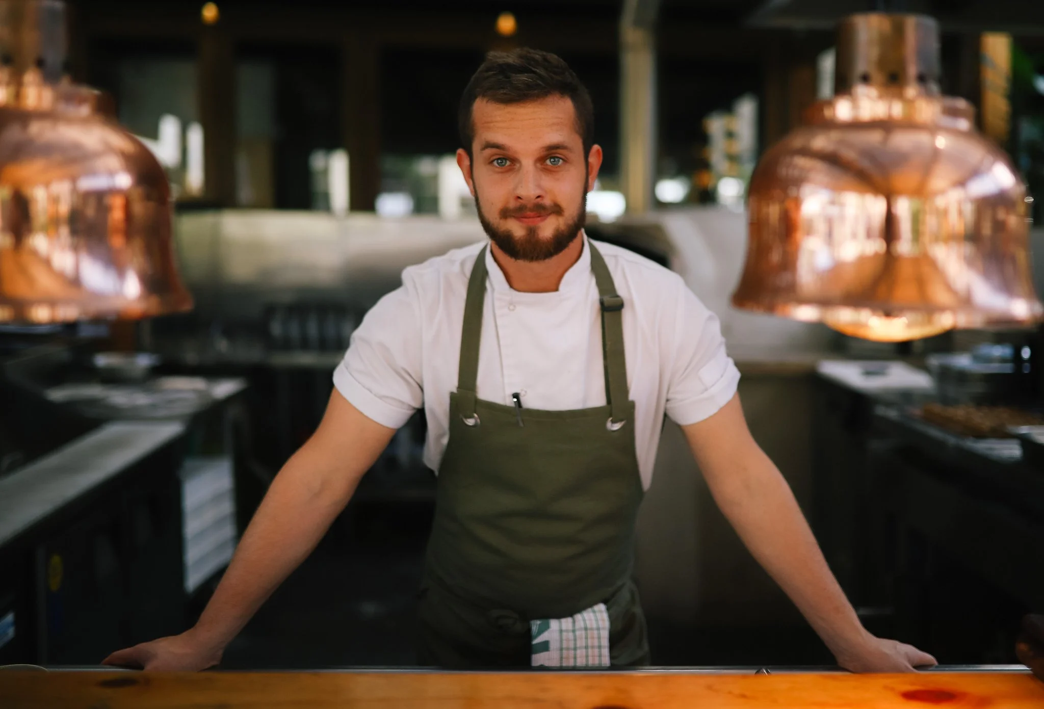 A male chef with a beard and blue eyes, wearing a white chef's coat and a green apron, standing behind a counter in a restaurant kitchen, looking at the camera with a slight smile. Copper pendant lights are hanging overhead.