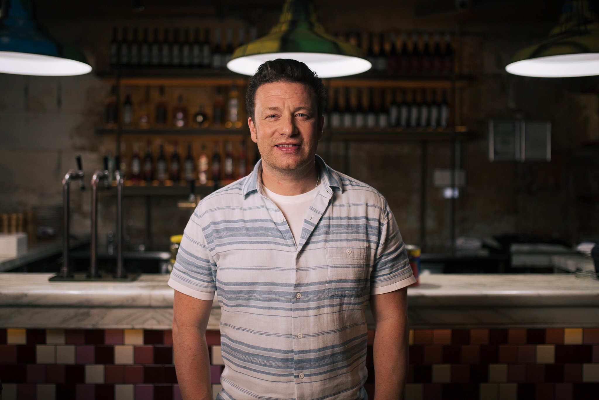 A man wearing a striped short-sleeve shirt standing in a bar or restaurant setting with liquor bottles on shelves behind him and green pendant lights overhead.