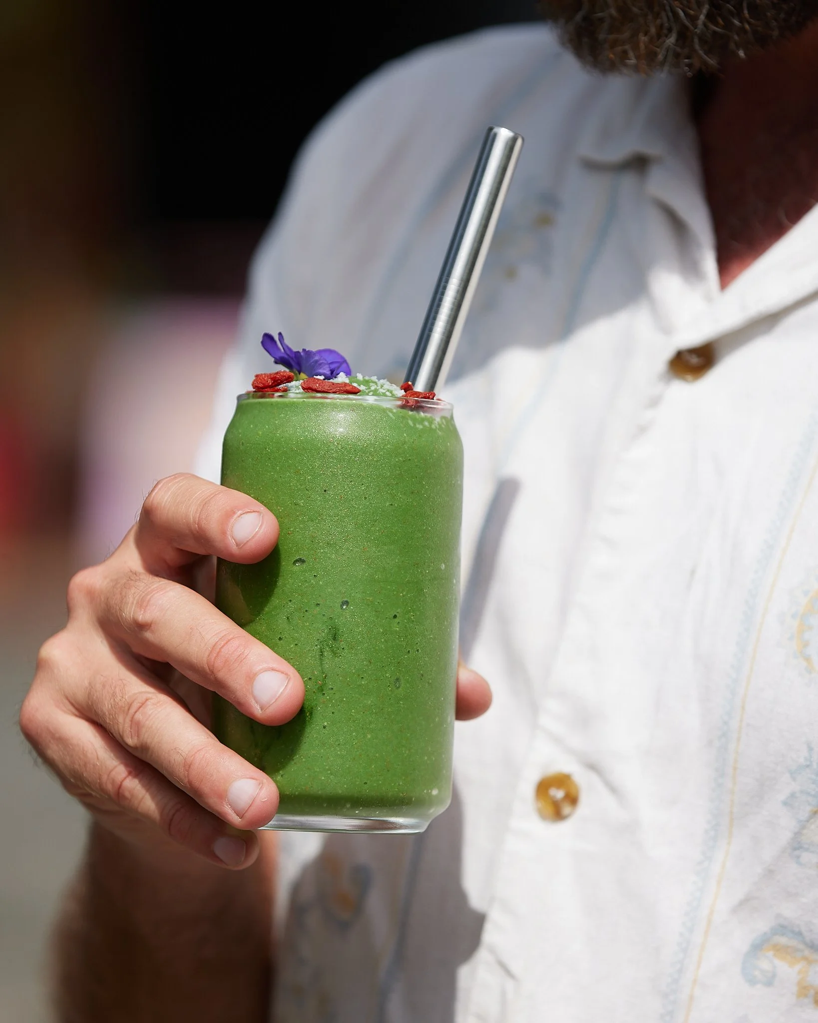 A person holding a green smoothie decorated with a purple edible flower, red goji berries, and white sesame seeds, with a metal straw.
