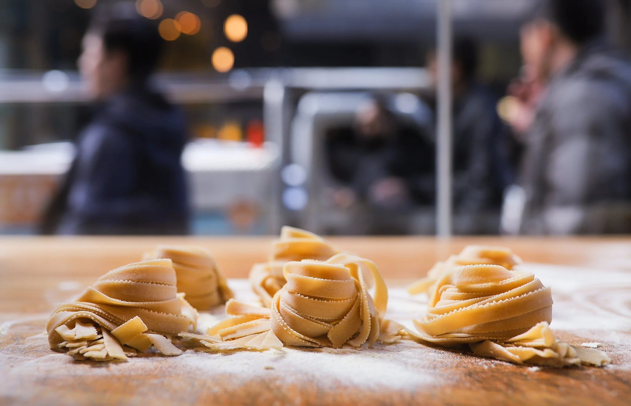 Fresh homemade pasta curls on a floured wooden surface with blurred people in a kitchen background.
