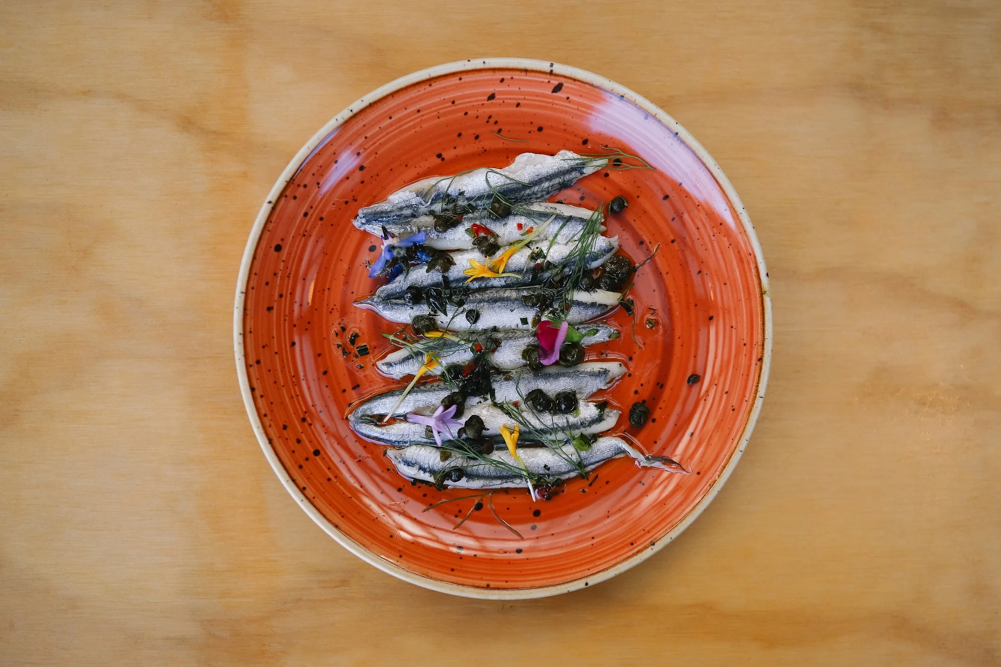 A plate of small fresh fish garnished with herbs, flowers, and black pepper on a red and white speckled ceramic plate on a wooden surface.