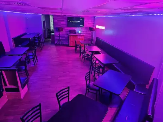 Empty small nightclub or karaoke room with purple ambient lighting, tables and chairs, a long bench along the wall, a stage with DJ equipment, and a large TV screen.