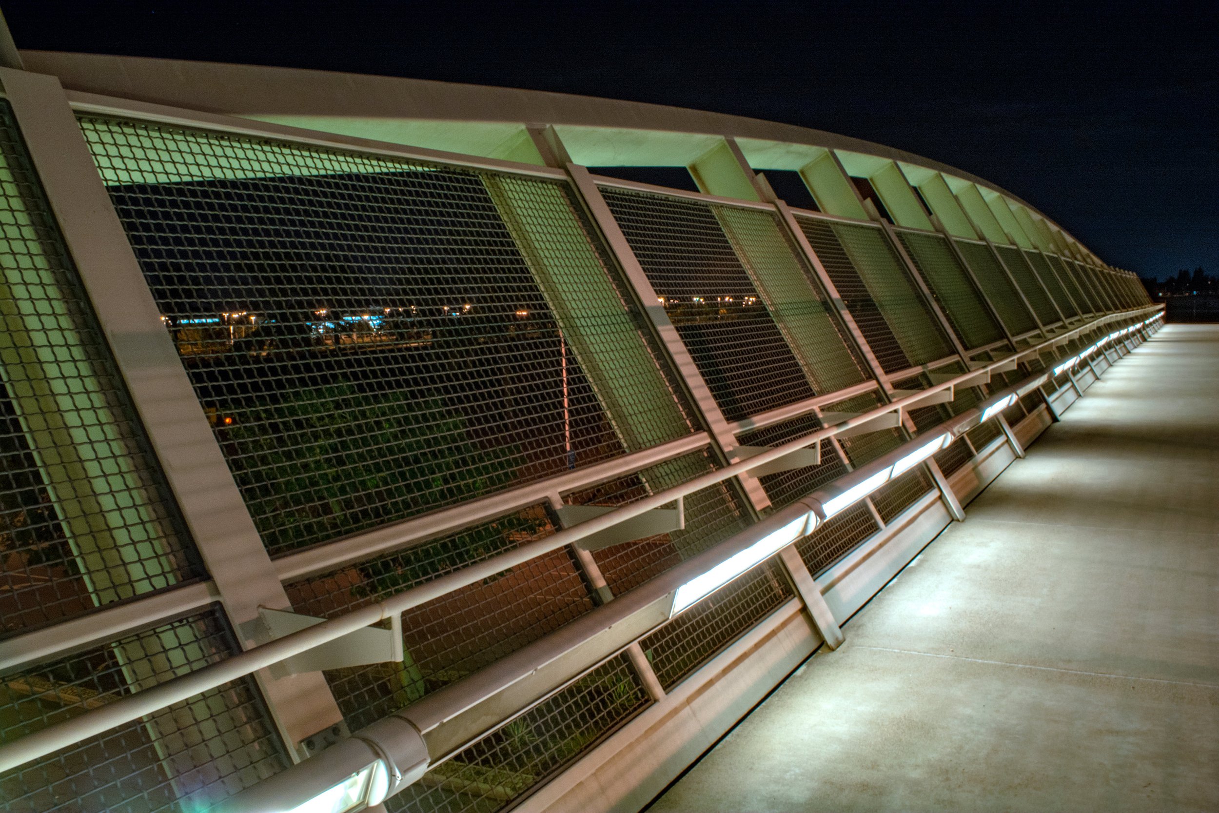 Night view of a modern pedestrian bridge with metal railings and mesh panels, illuminated by strip lights along the walkway.