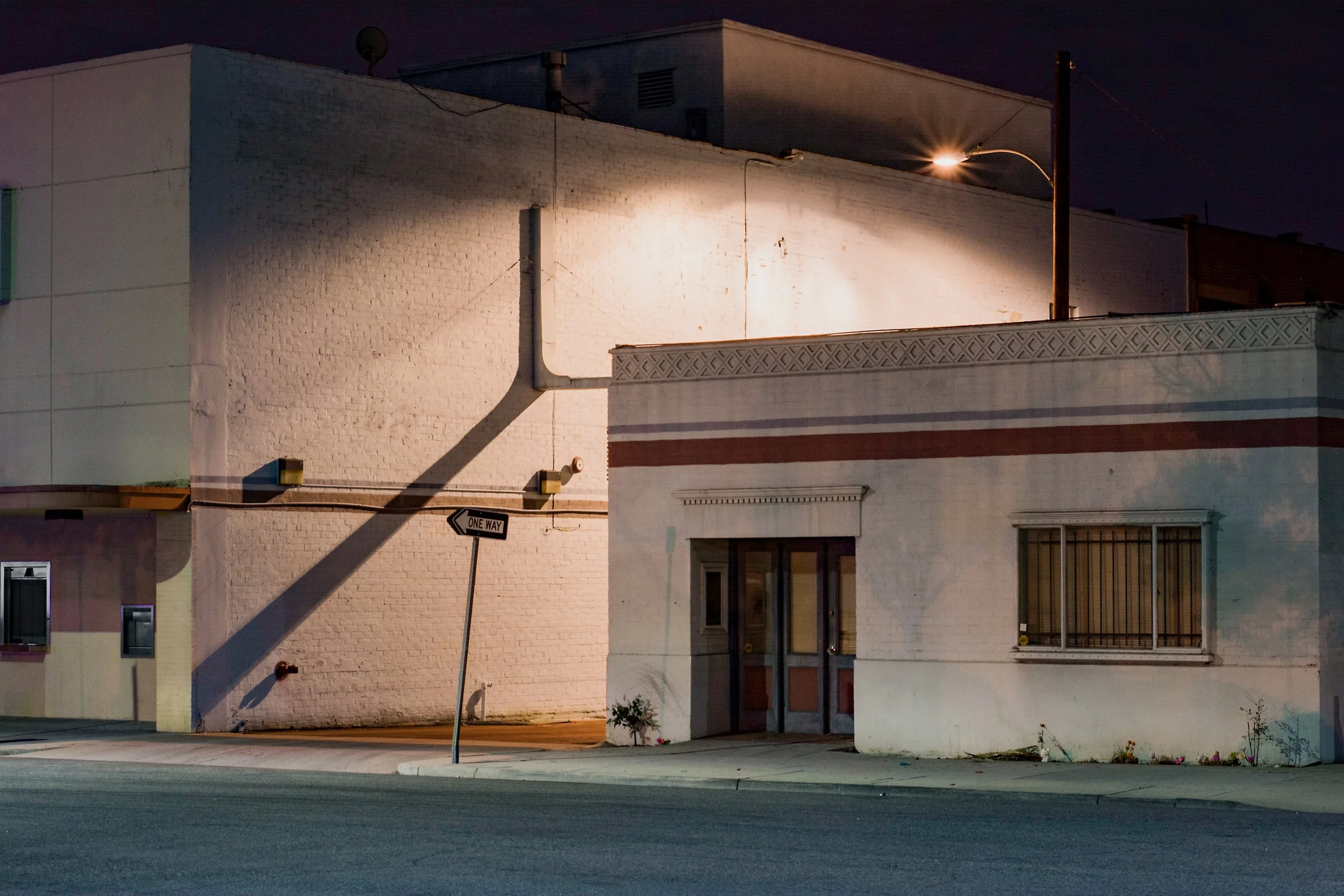 Nighttime street view of a white brick building with two entrances, one with a door and the other with barred window, illuminated by a streetlight with a one-way street sign in the foreground.