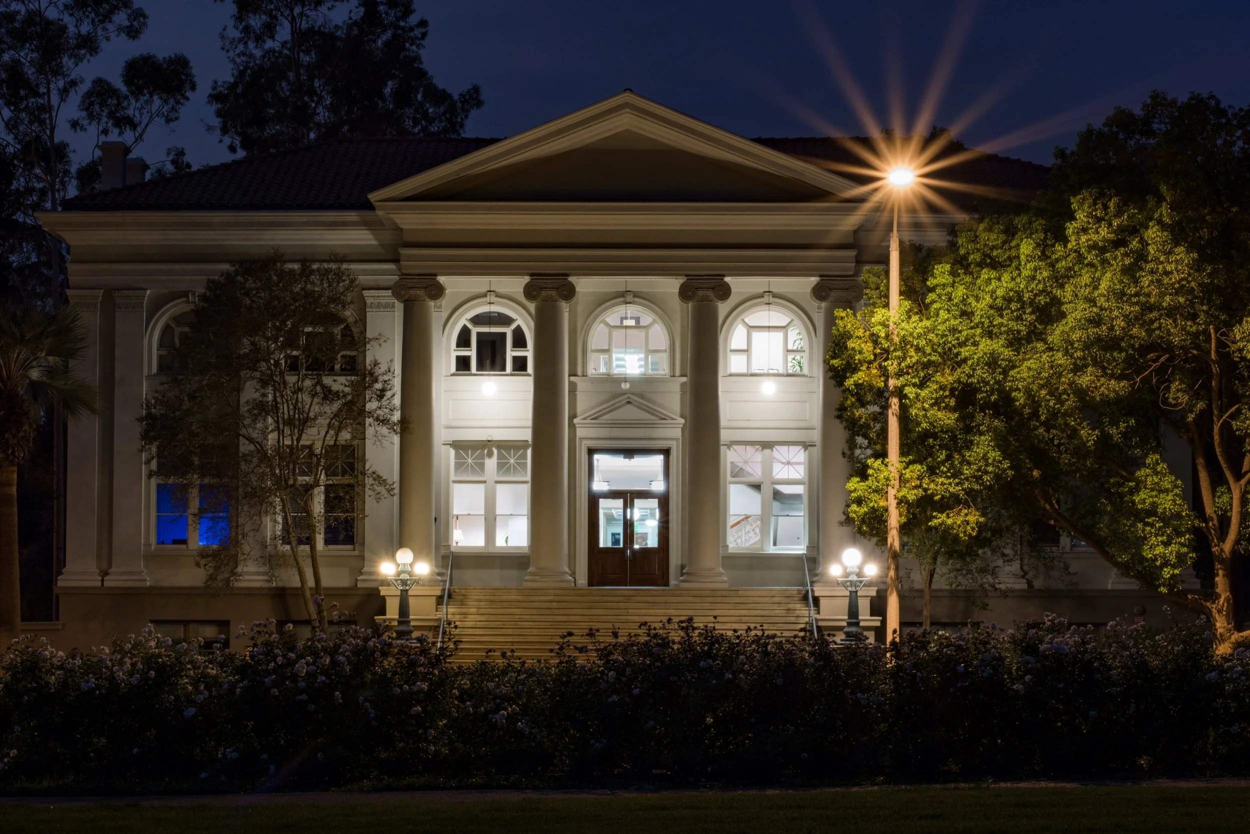 Beautifull historic building at night with illuminated windows and exterior lights, surrounded by trees and bushes.