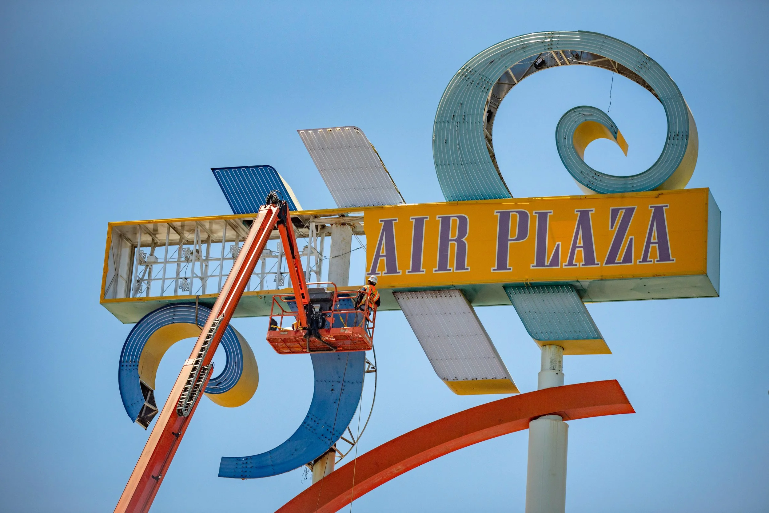 Worker on a cherry picker repairing or maintaining the vintage Air Plaza sign against a clear blue sky.