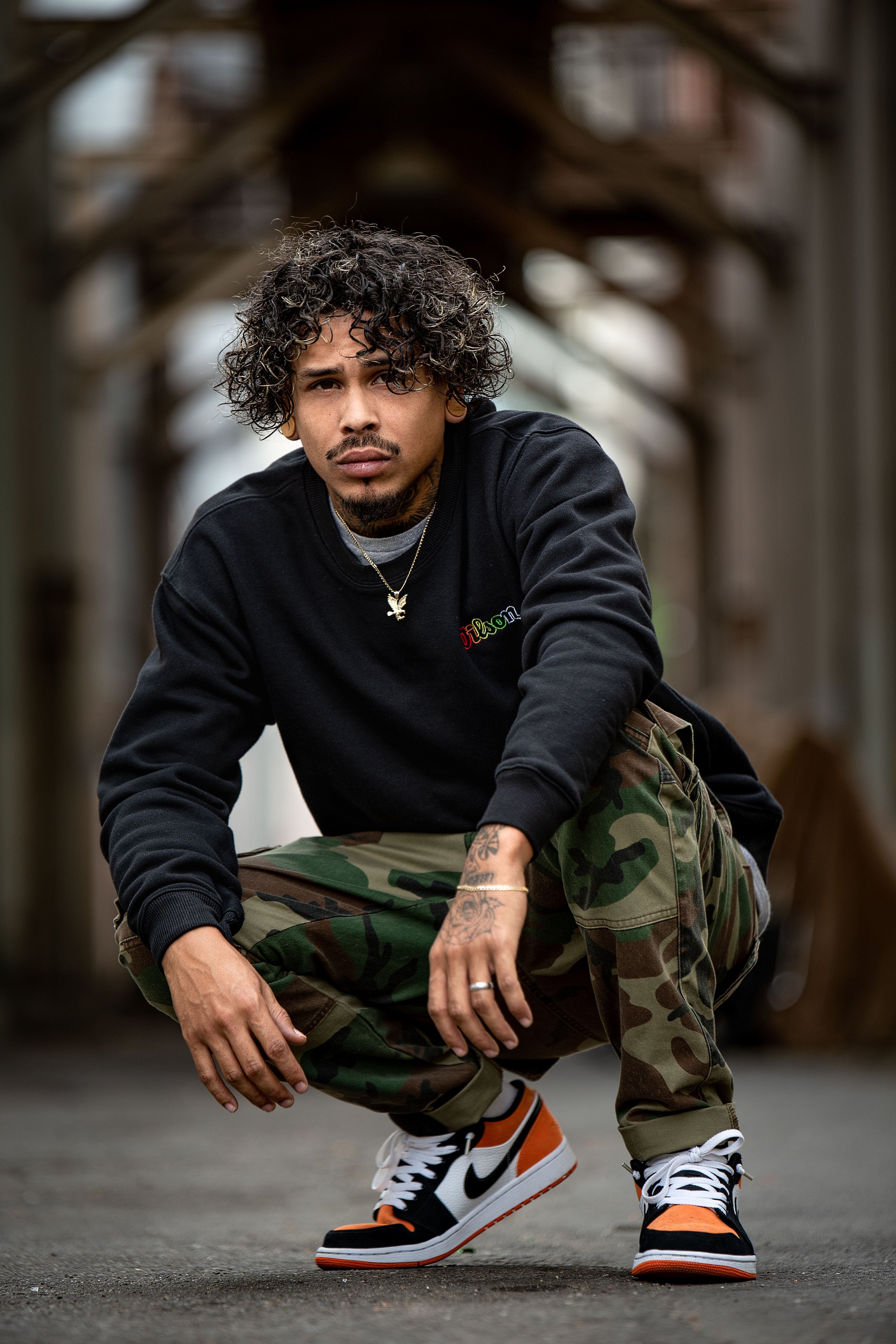 A young man with curly hair and tattoos crouching outdoors on a paved surface, wearing a black sweatshirt, camouflage pants, and Nike sneakers, with a blurred urban background.