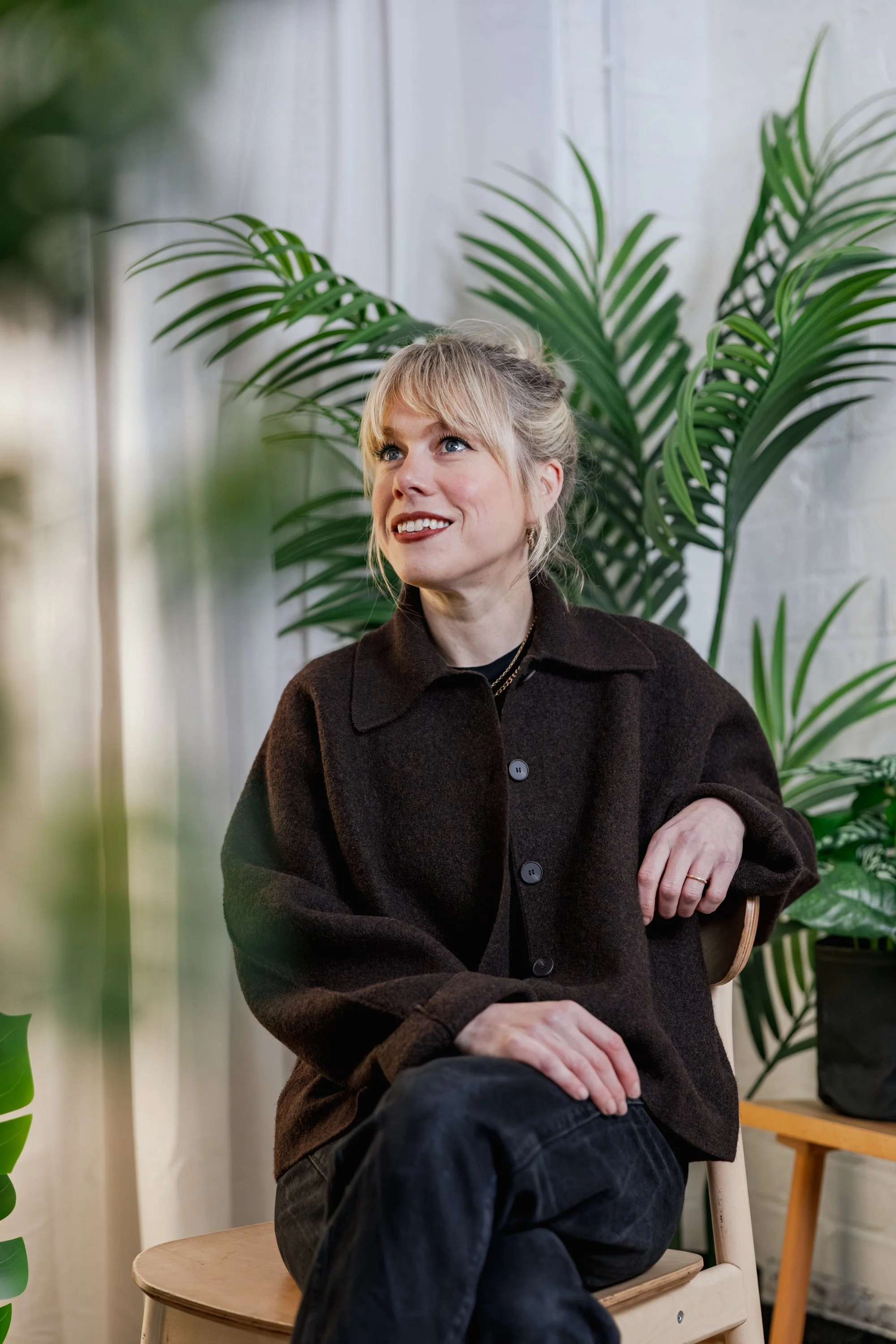 Portrait of a blonde woman with a fringe sitting on a wooden chair in a studio at GRIT, Stockport, smiling and looking off camera, surrounded by lush green plants and soft natural light against a white brick wall.