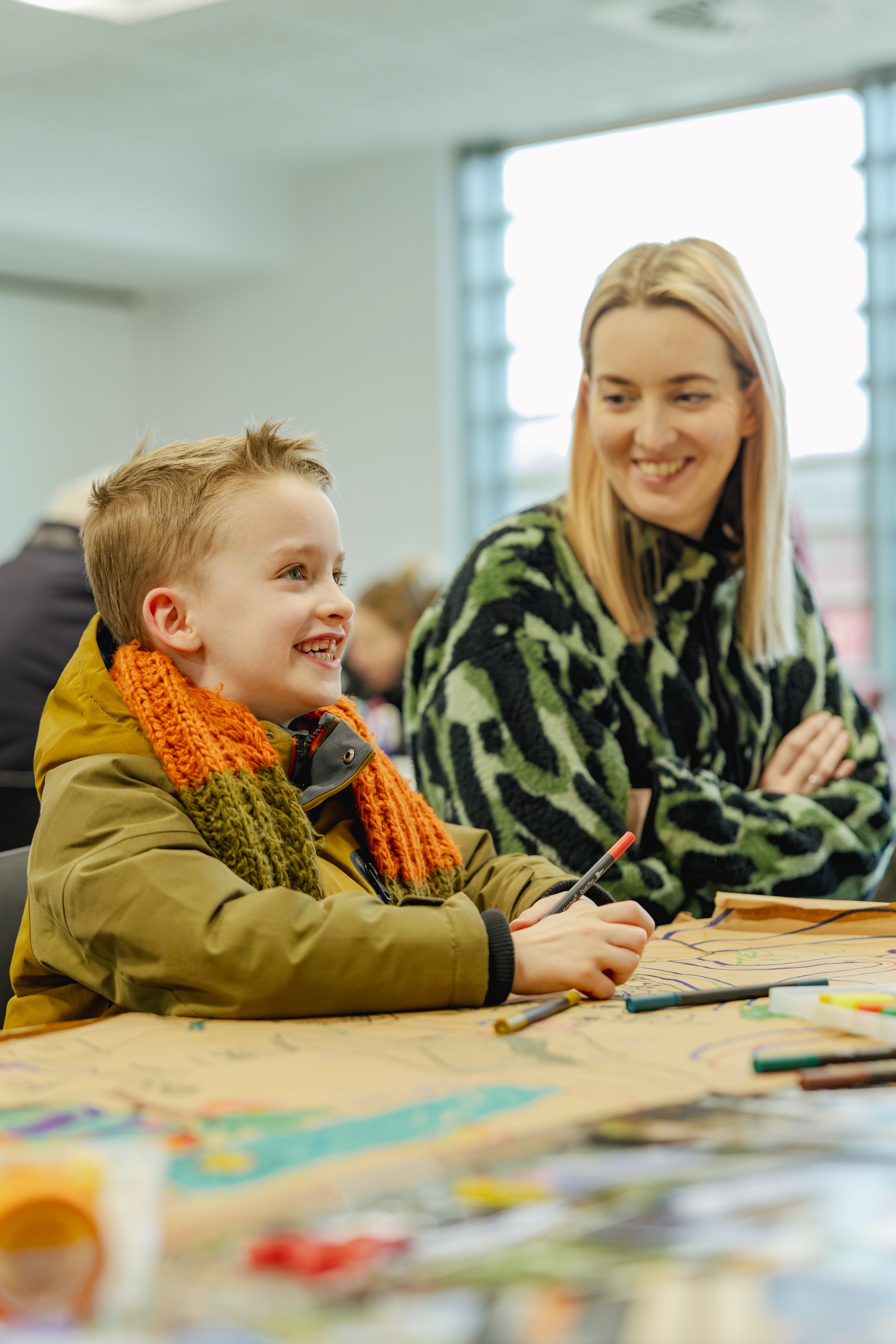 Candid workshop photography of a child smiling while drawing at a creative learning session, with a facilitator engaging in the background in a bright workshop environment.
