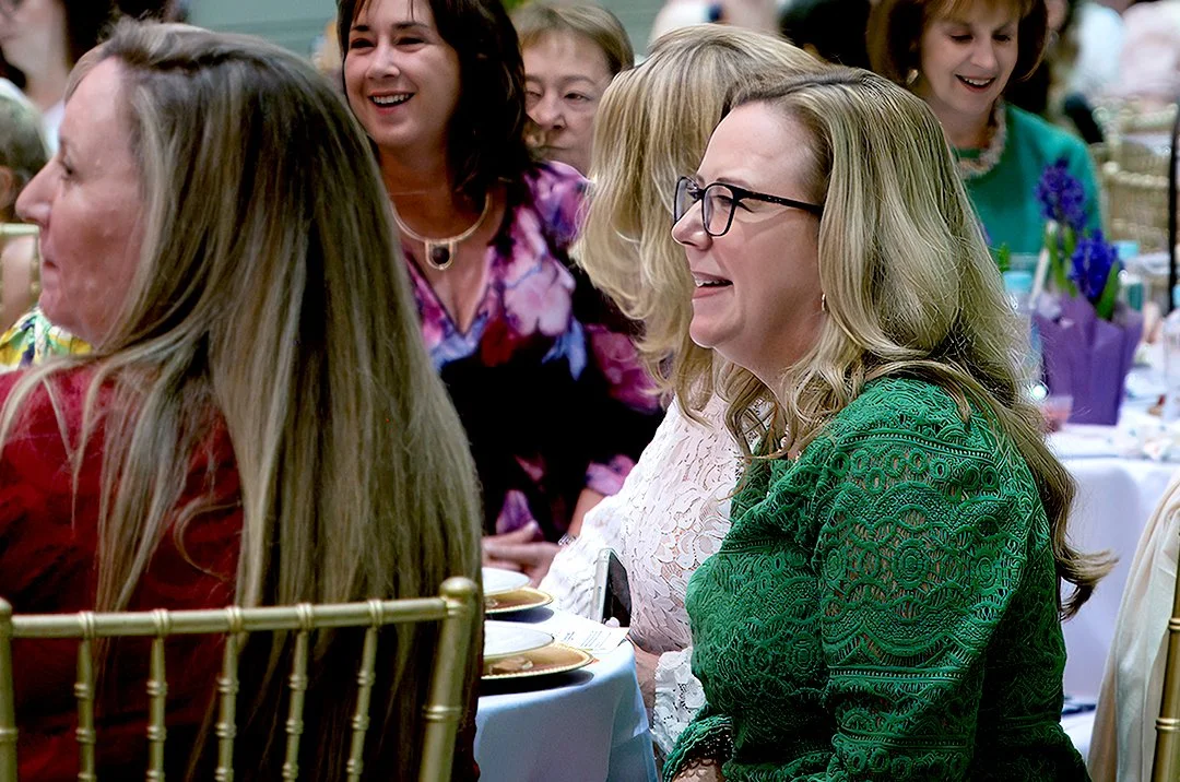 Smiling women seated at a formal event, dressed in colorful attire and surrounded by decorated tables.