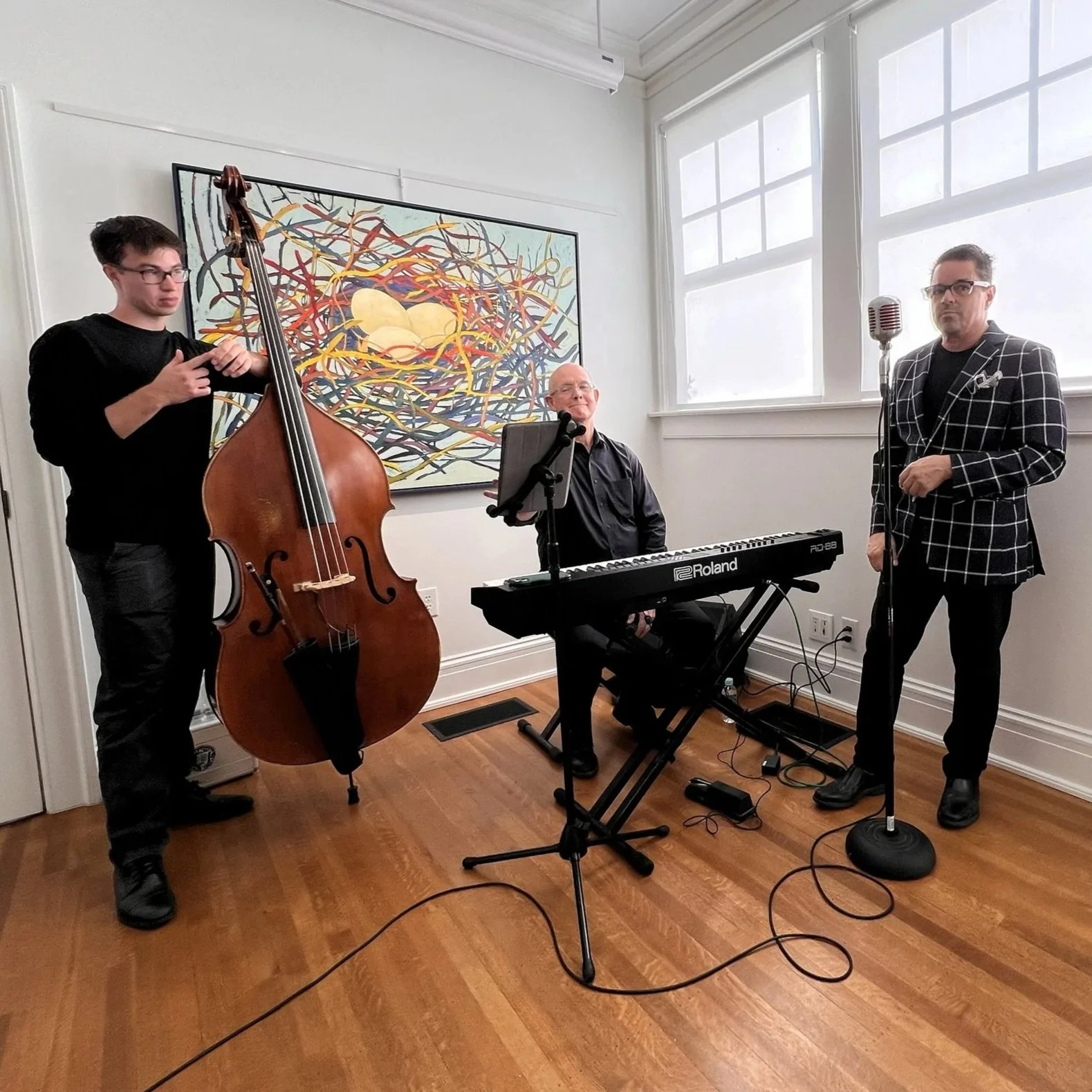 Three musicians with a double bass, keyboard, and microphone in a room with wooden floors and white walls, featuring abstract art on the wall and large windows.