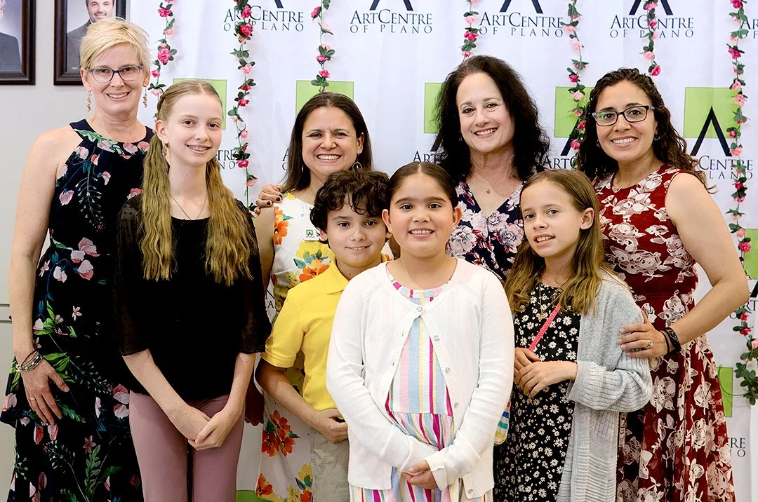 A group of people, including adults and children, pose together in front of a banner for the ArtCentre of Plano. The banner is decorated with floral designs. The group is dressed in a variety of colorful outfits.