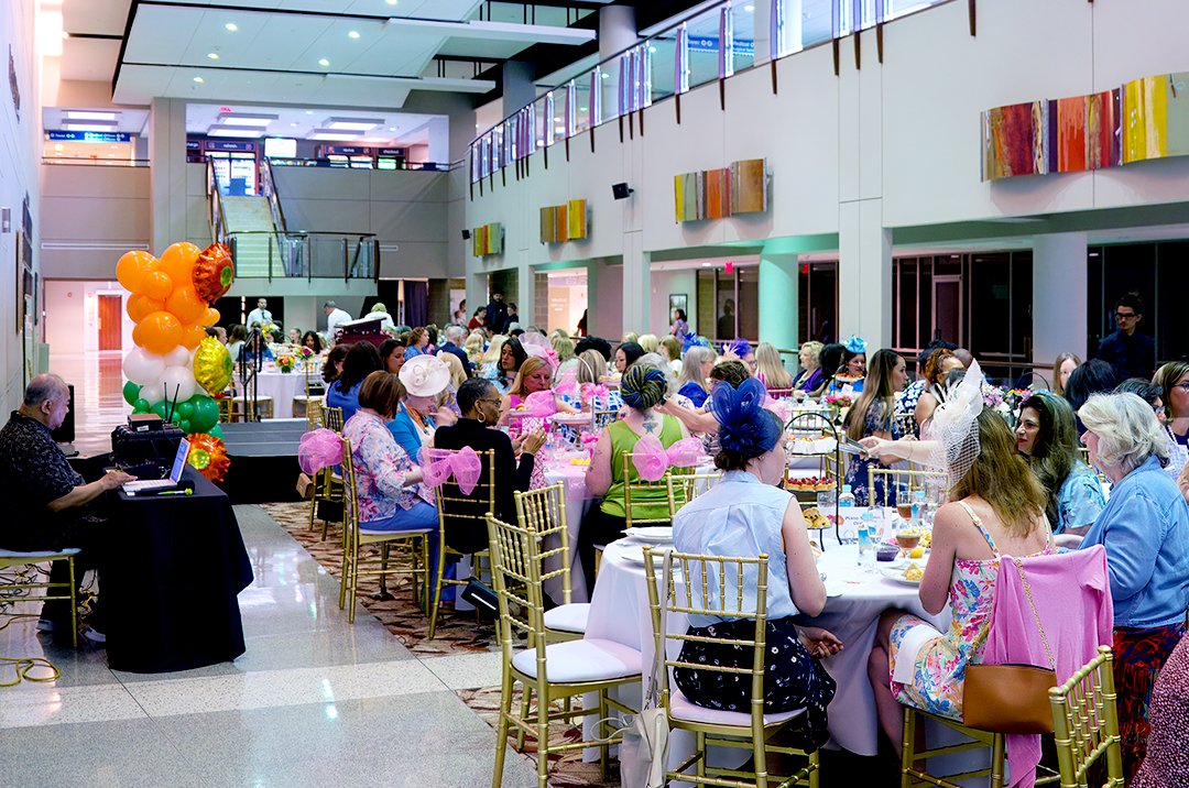 A large group of people seated at decorated tables in a spacious hall, possibly attending a formal event. The attendees wear colorful attire and hats, and there are flower and balloon arrangements nearby.