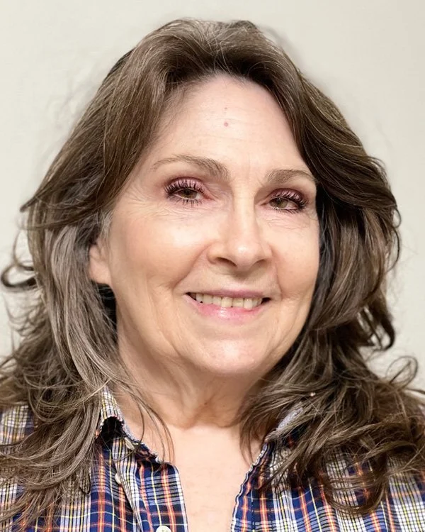 Close-up of a smiling woman with long, wavy brown hair, wearing a plaid shirt, against a plain light-colored background.