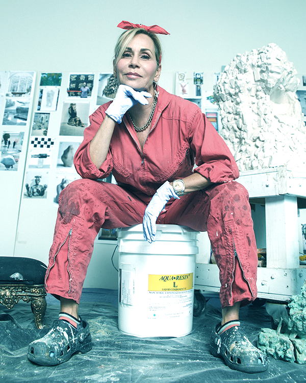 A woman wearing a red jumpsuit, gloves, and a red headband sits on a bucket in an artist's studio, surrounded by art supplies, sculptures, and photos on the wall.