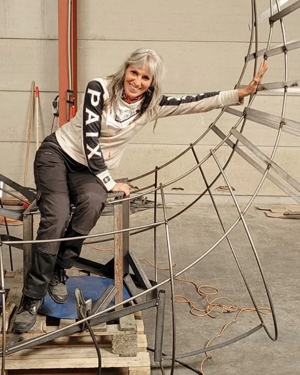 A woman smiling and crouching on a metal scaffolding structure in an industrial workshop or warehouse, holding onto the frame of what appears to be a large, curved metal framework or sculpture.