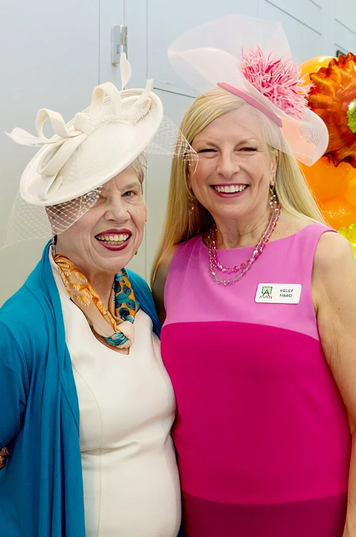 Two women smiling, wearing elegant hats, one in blue and white attire with a colorful scarf, the other in a pink dress with a name tag.