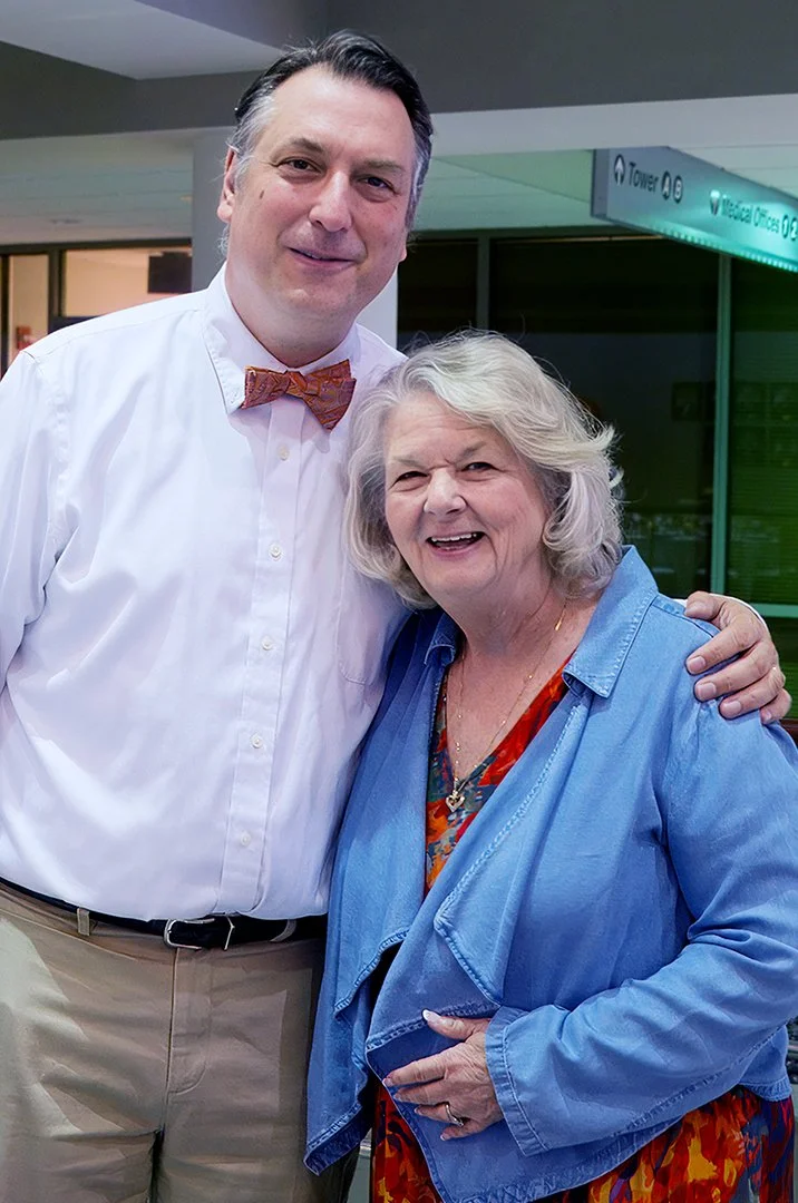 A man wearing a white shirt and bow tie stands next to an older woman in a blue jacket. They are smiling and posing together indoors.