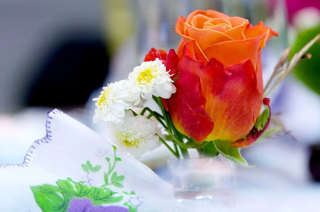 Close-up of an orange and red rose with small white flowers in a clear vase next to a white cloth with green and purple floral embroidery.