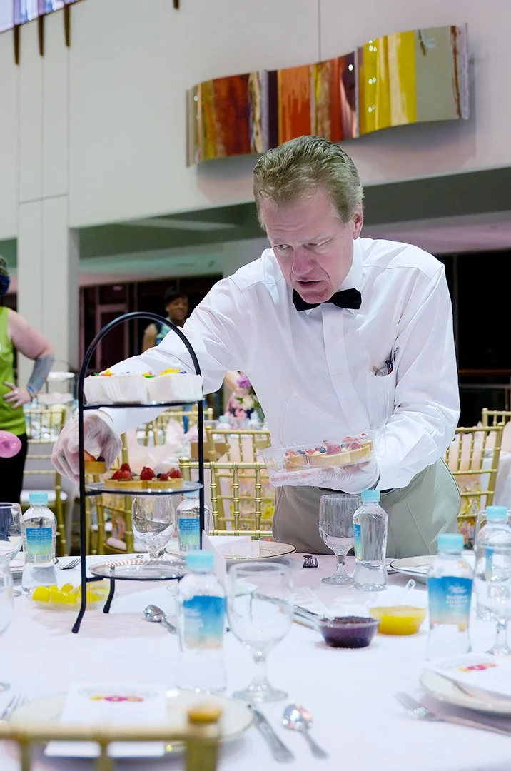 Man arranging desserts on a table at an event, wearing a white shirt with a bow tie.