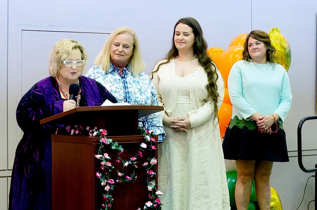Four women standing at a podium with floral decoration, holding a microphone, and smiling. Orange and green balloons in the background. Casual and formal attire visible.