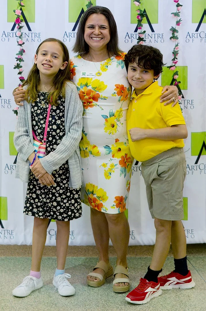 A woman in a floral dress is smiling with her arms around two children, one wearing a black floral dress and the other in a yellow shirt. They are at the ArtCentre of Plano.