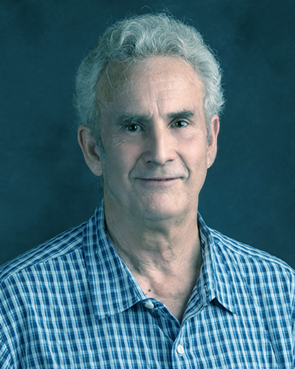 Portrait of an older man with white curly hair, wearing a blue checkered shirt, standing against a dark background.