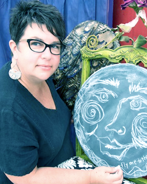 Woman with short dark hair and glasses holding a chalkboard art piece with a face drawing and the words 'fly me to the' visible. She is wearing earrings and a dark top, with colorful artistic objects and flowers in the background.