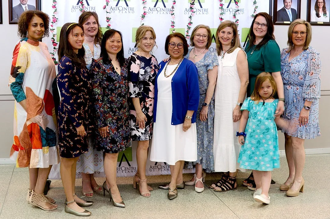 A group of women and a young girl posing together in front of a backdrop labeled 'Art Centre of Plano.' The women are wearing a variety of colorful dresses, and the young girl is in a turquoise dress. They are standing indoors in a brightly lit envir