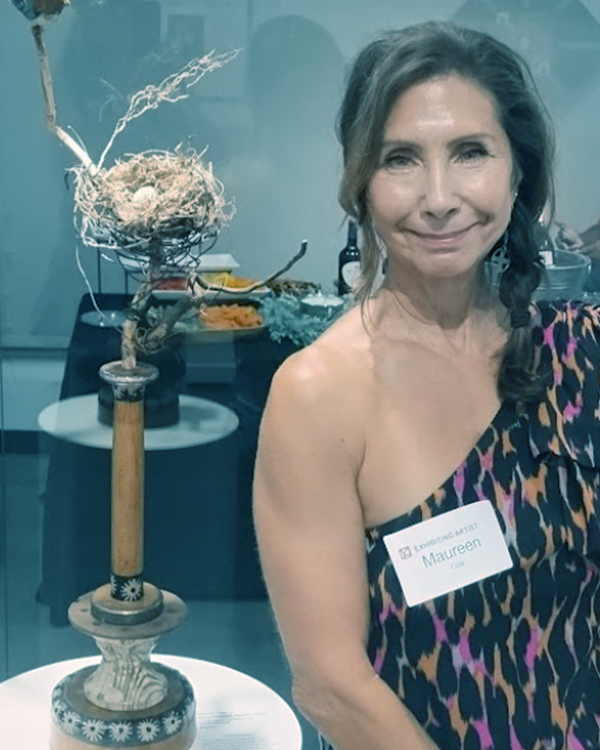 A woman with long wavy hair, wearing a one-shoulder leopard print dress, smiling at the camera, standing next to a tall, decorative lamp on a table, with a name tag that reads 'Maureen'.