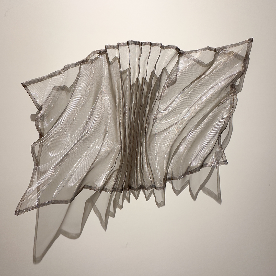 An abstract sculpture made of layered, pleated, and semi-transparent fabric or material, creating a ruffled, floral-like shape with shadow cast on a plain wall.