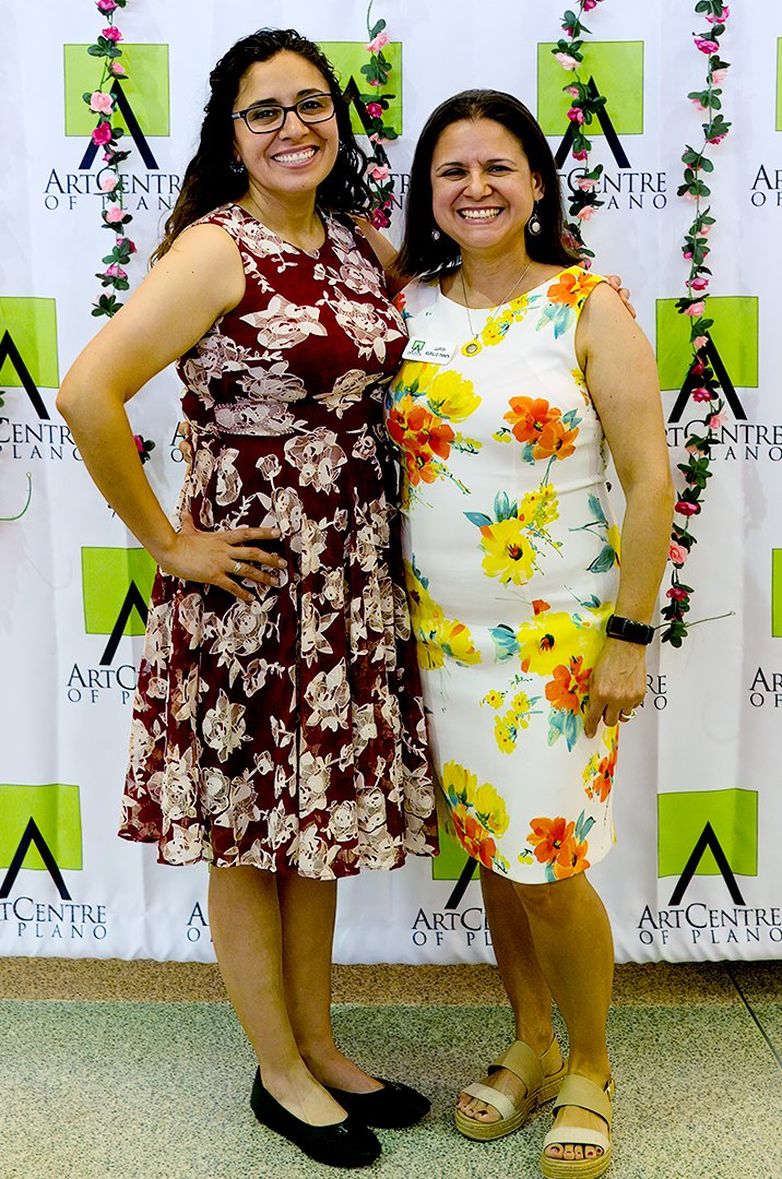 Two women standing together, smiling, in front of an Art Centre of Plano backdrop. One is wearing a maroon dress with floral patterns, the other a white dress with colorful flowers.