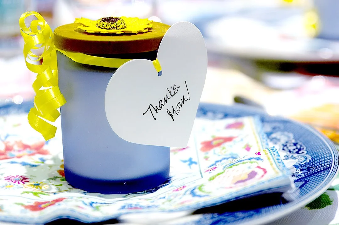 Glass jar with a wooden lid, yellow ribbon, flower decoration, and a heart-shaped tag reading 'Thanks Mom!' on floral napkins.