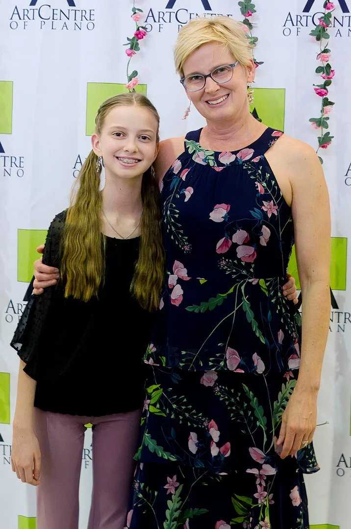 Two people smiling in front of a backdrop with "Art Centre of Plano" logos; one is wearing a floral dress and glasses, the other a black top and pink pants.