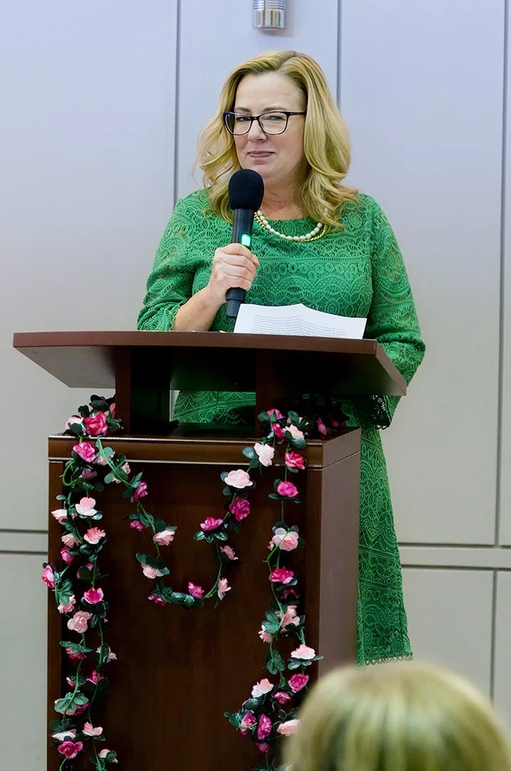 Woman in a green dress speaking at a podium adorned with flowers, holding a microphone and papers.