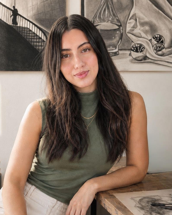 A young woman with long, dark, wavy hair sitting at a wooden table indoors, wearing a sleeveless green top and layered necklaces, looking at the camera with a slight smile. There is a framed black-and-white artwork of a staircase and abstract objects hanging on the wall behind her.