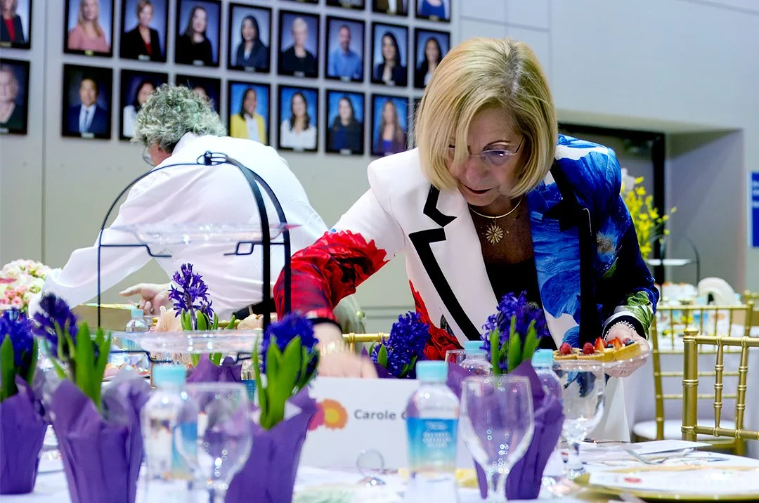Woman arranging table with cupcakes and purple flowers; wall of portraits in background.