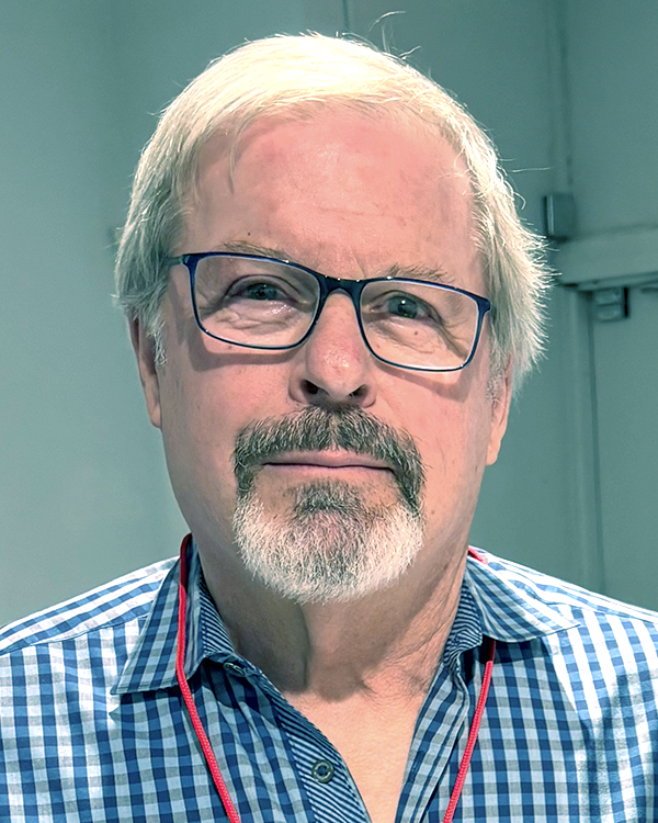 A middle-aged man with blond hair, glasses, and a beard and mustache, wearing a blue checkered shirt and a red lanyard, standing indoors in front of a gray wall.