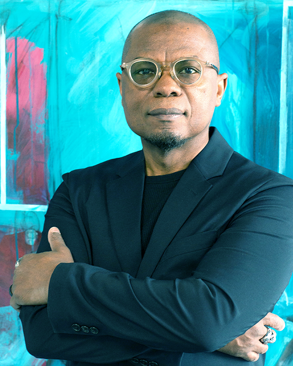 A man with glasses and a shaved head wearing a black blazer, standing with arms crossed in front of a colorful abstract painting background.