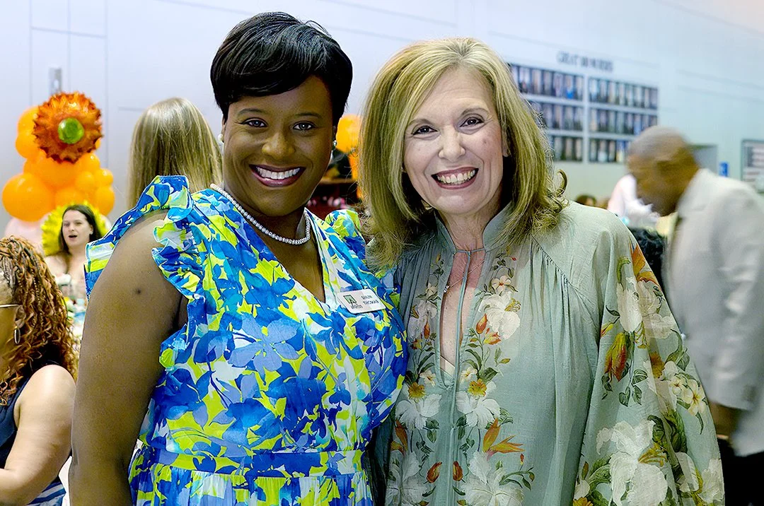 Two smiling women pose together indoors, surrounded by other people and orange balloon decorations.