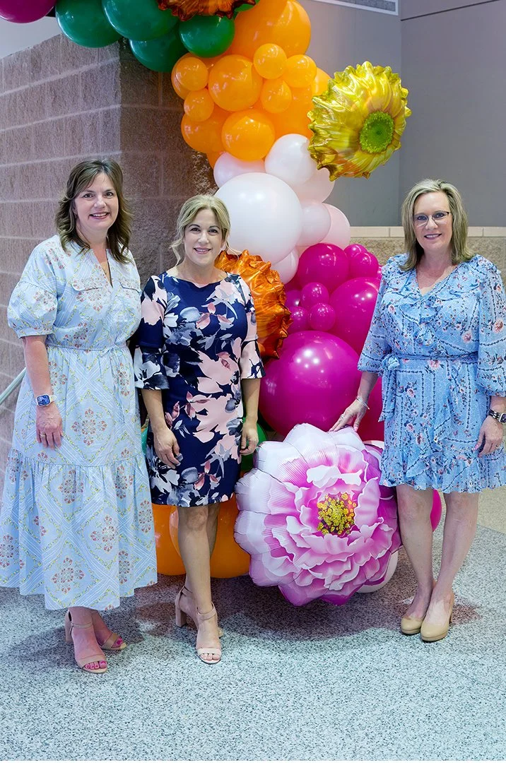 Three women in floral dresses standing near colorful balloon display and large flower decorations indoors.