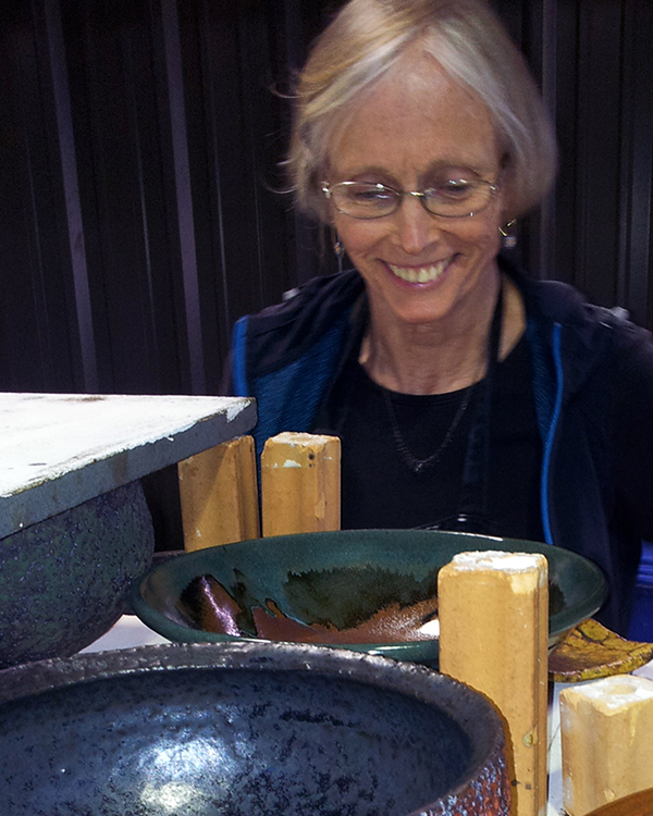 An elderly woman with short gray hair and glasses smiling while looking at empty pottery bowls on a table.