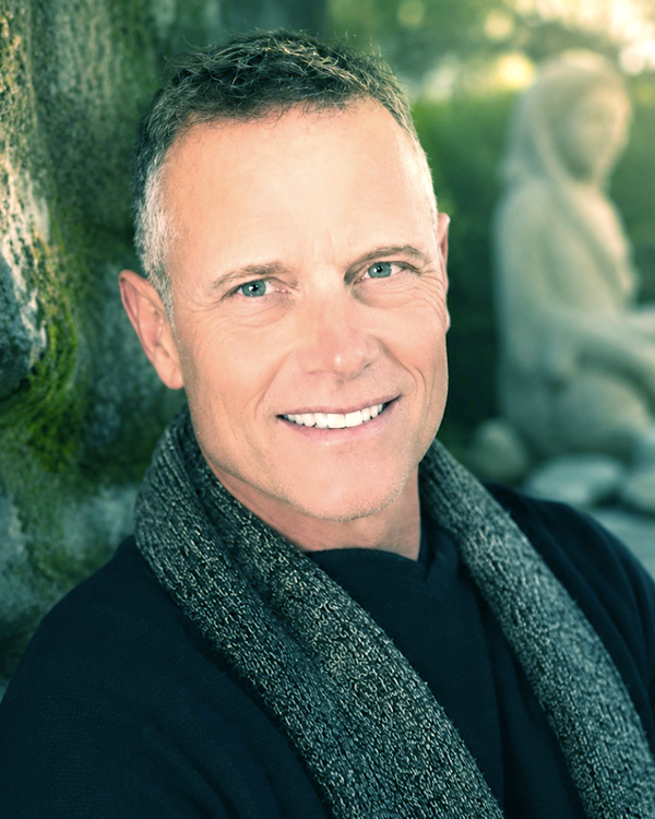 A smiling middle-aged man with short, gray hair and blue eyes, wearing a black shirt and a textured gray scarf, stands outdoors near a moss-covered tree with a blurred statue in the background.
