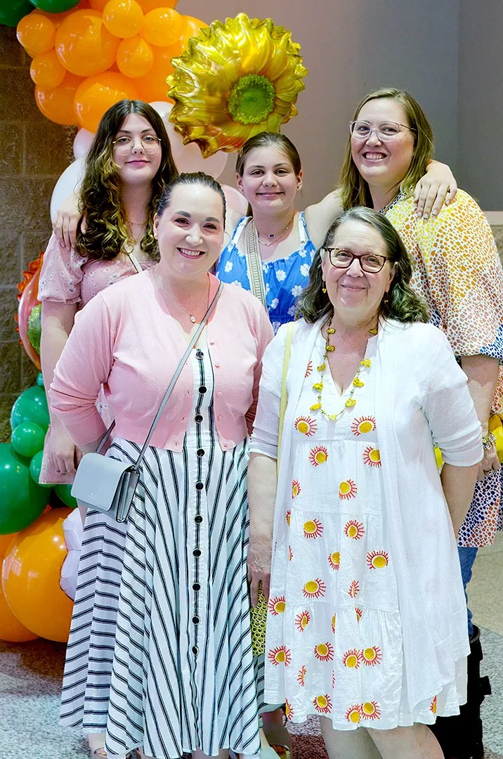 Group of five women smiling in front of colorful balloon decoration with sunflower design.