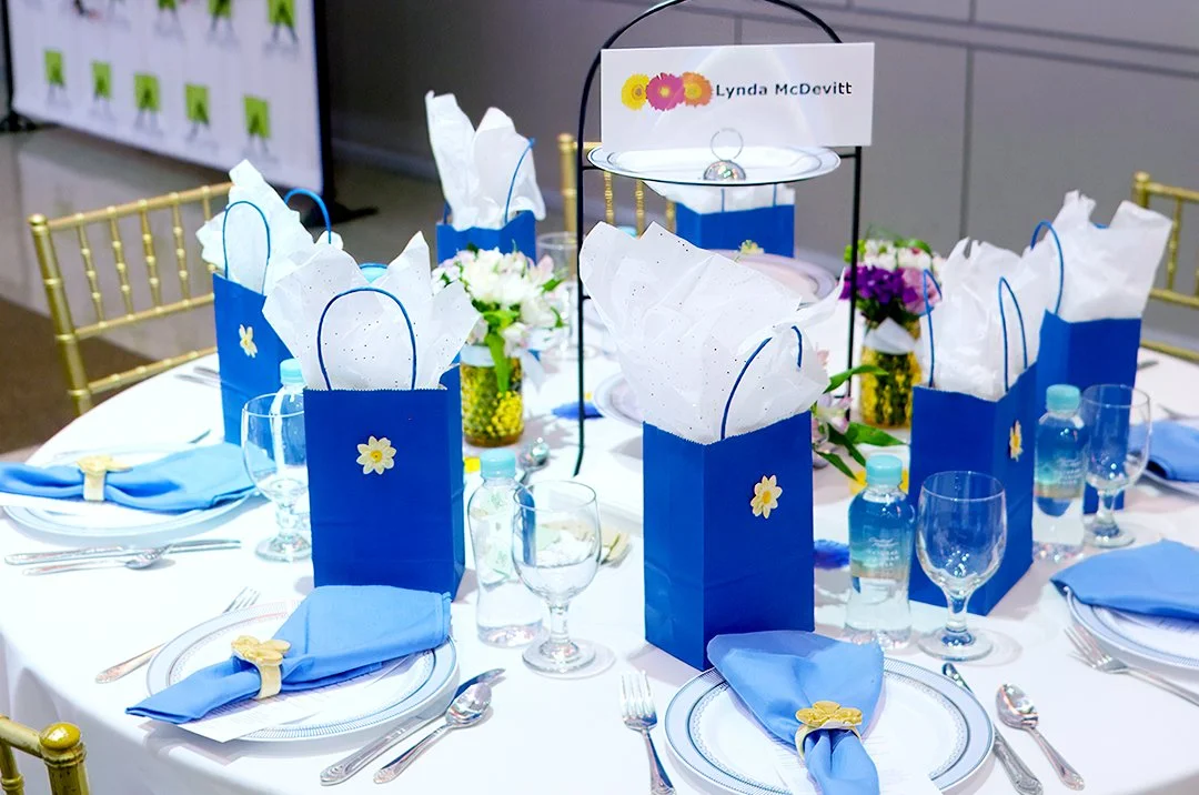 Elegant table setting with blue gift bags, white tissue paper, blue napkins, and floral decorations.