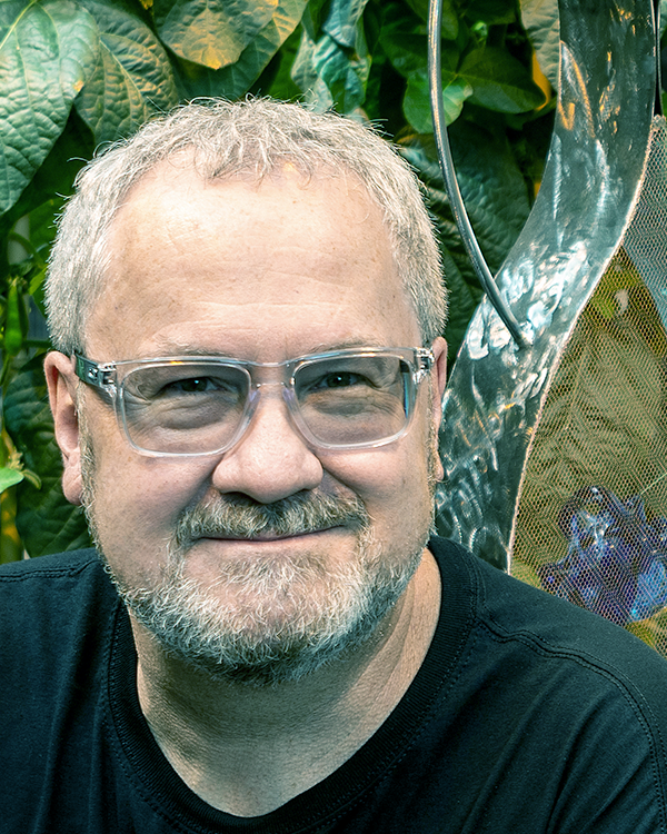A middle-aged man with short gray hair, glasses, and a beard, smiling in front of a leafy green background.