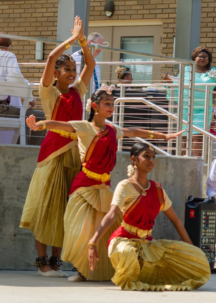 Three young women performing a traditional dance in colorful attire, with gold and red outfits, in an outdoor setting. They are positioned near a building with onlookers in the background.