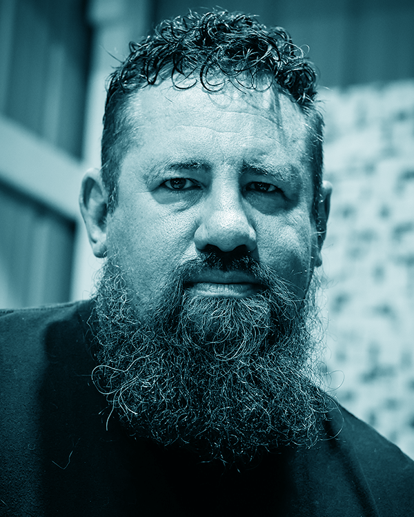 Close-up of a man with curly hair and a full beard, wearing a dark shirt, in an indoor setting.
